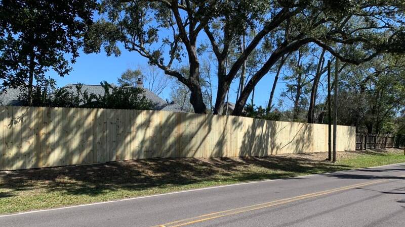 Long wood privacy fence with vertical boards along a two-lane road under live oaks