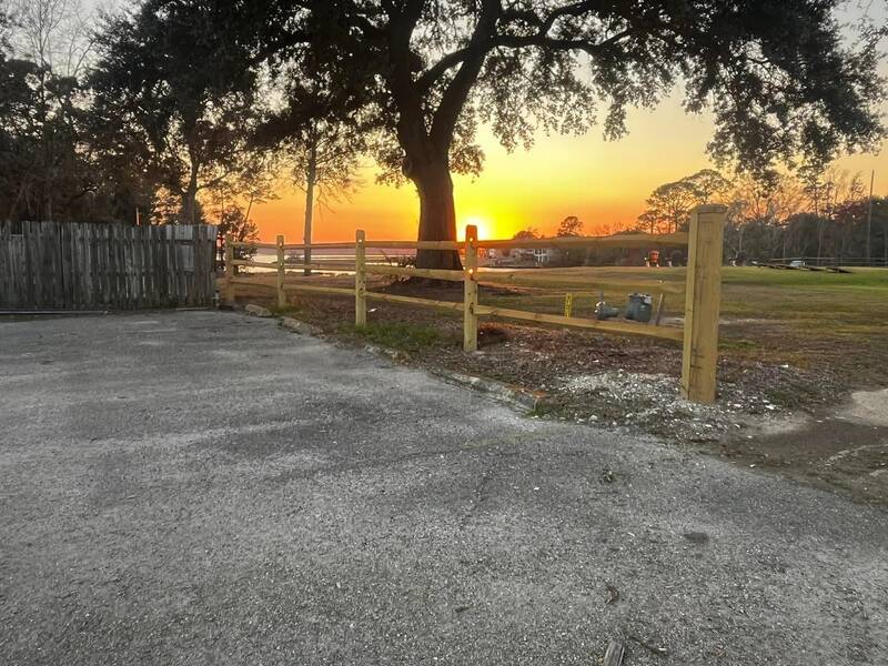 Wood post-and-rail fence along a paved lot at sunset with a live oak behind